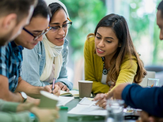 Five students working together at a table with pen and paper.